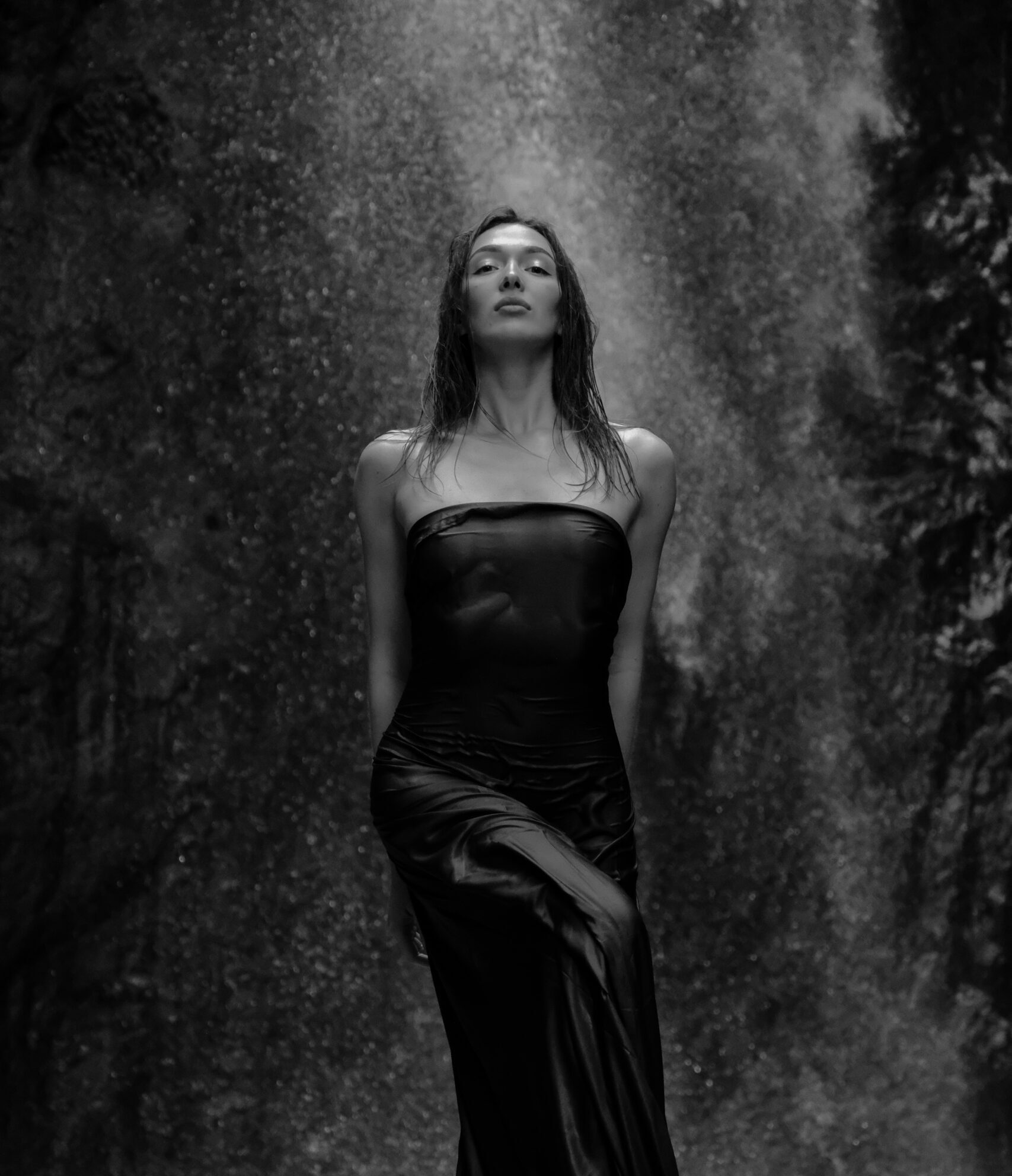 Black-and-white editorial portrait of Yuliya standing beneath a waterfall in a long dark dress.
